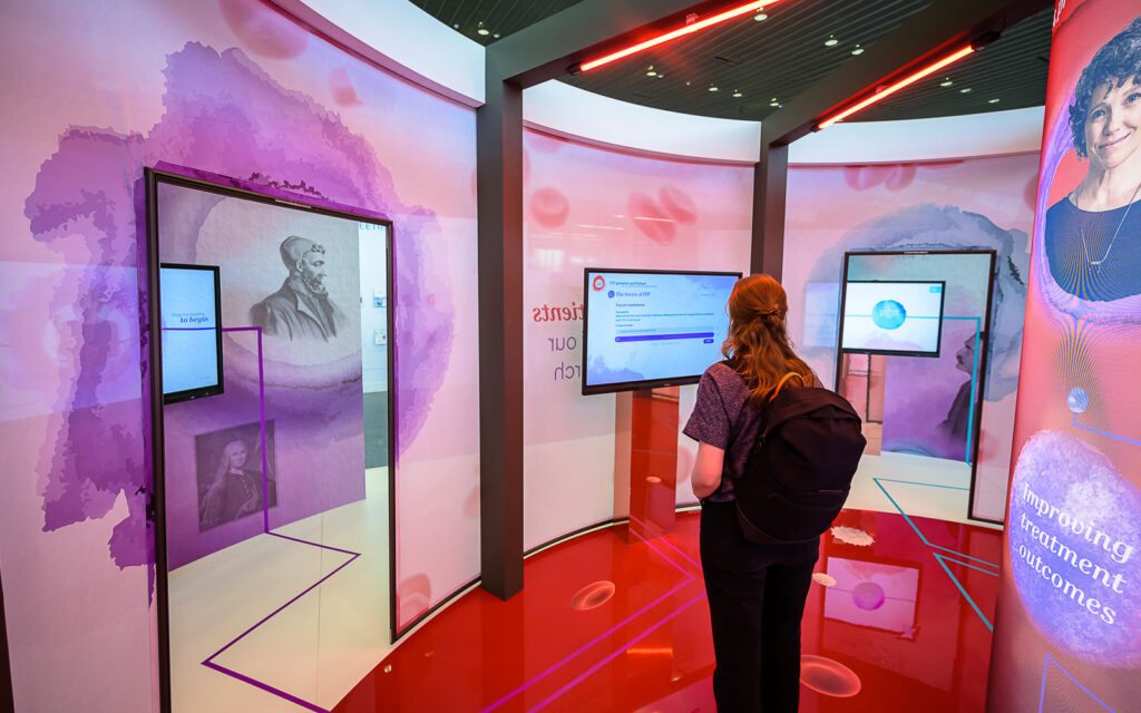 A Congress attendee in the EHA2023 exhibition space. Around her are screens displaying information and illustrations of blood cells.