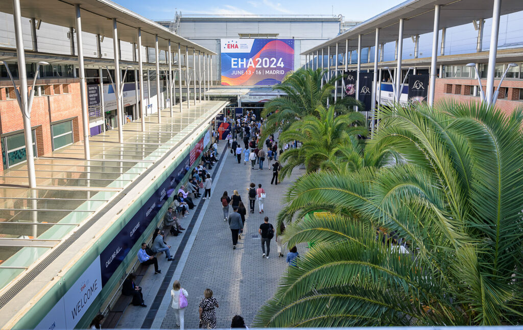 The EHA2024 venue at IFEMA, Madrid, Spain, shot from an upper floor. Below, attendees are walking around an uncovered space. There are around four palm trees in front of the camera.