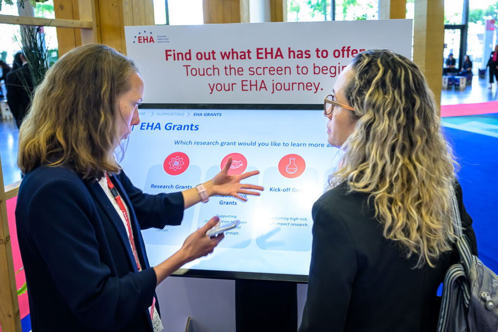 Two women interacting with a touchscreen display. The display has 'EHA Grants' at the top, and options for 'Research Grants,' 'Collaborative Grants,' and 'Kick-off Grants.'