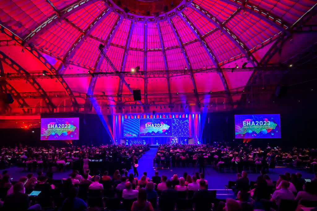 A wide shot of a large hall at the EHA2023 venue. Hundreds of attendees are seated facing a stage. Three large screens display 'EHA2023.'
