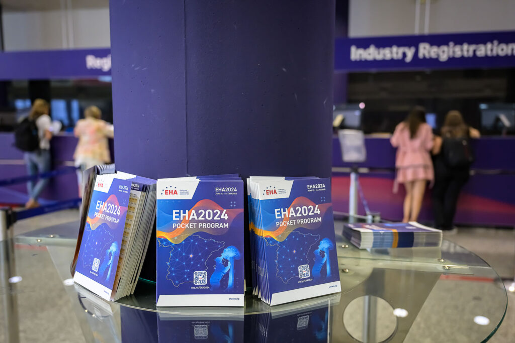 Copies of the EHA2024 pocket program on a glass table. The programs are leaning against a column that runs through the center of the table.
