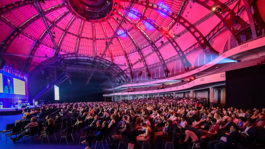 Hundreds of EHA2023 attendees watching a presentation in a large hall. The space has dome-shaped ceiling that is lit in red.