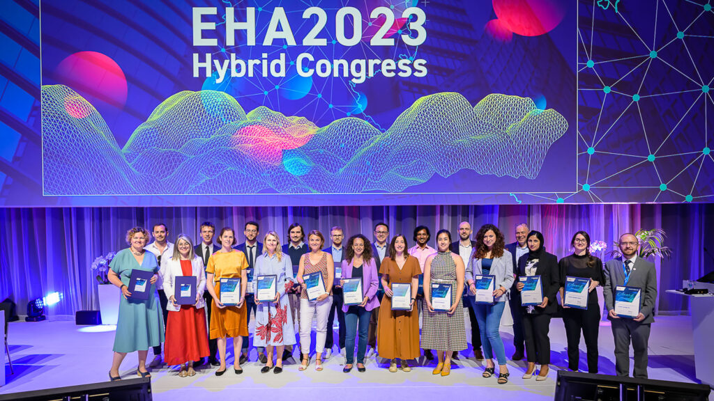 Around 20 EHA award winners on stage. Behind them is screen displaying the words "EHA2023 Hybrid Congress."