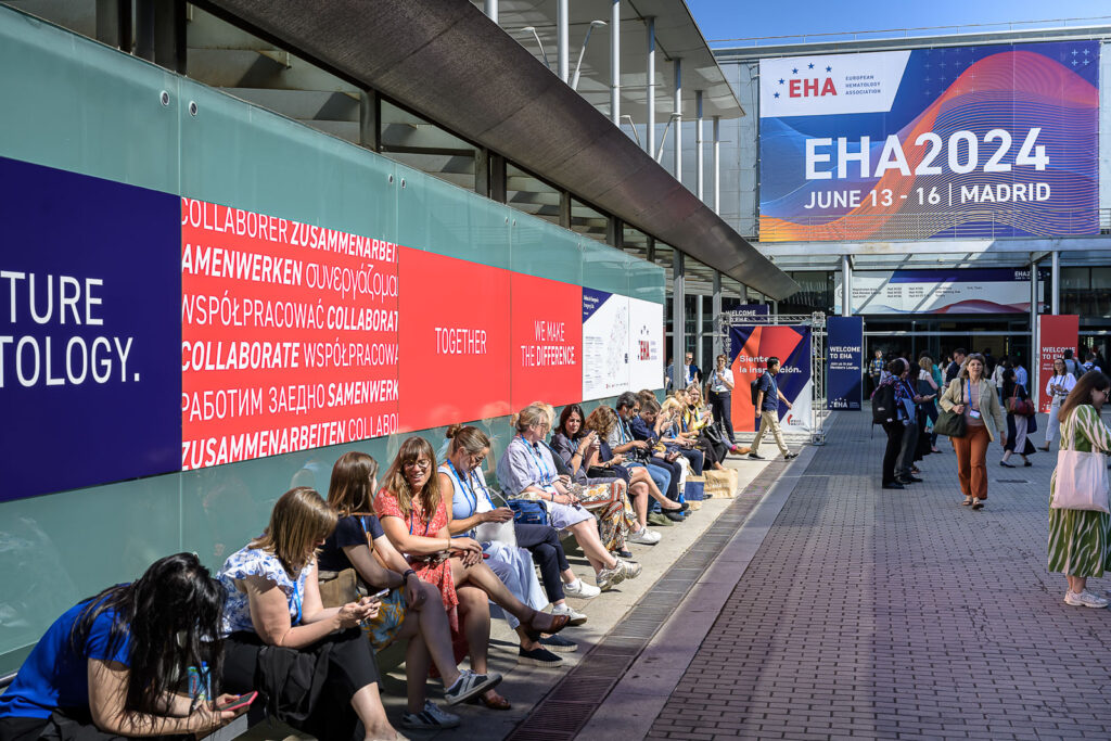 A busy outdoor space in the EHA2024 venue. Many attendees are seated and have their backs to a frosted glass wall. Around them are signs promoting EHA.