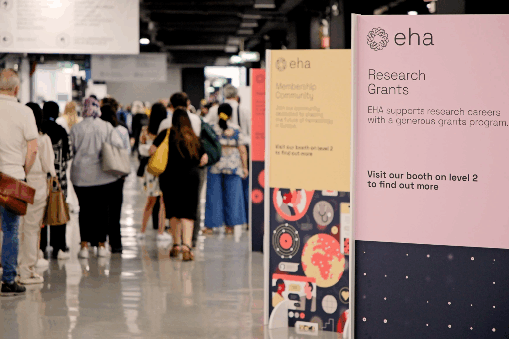 A small number of delegates walking down one of the main corridors of the EHA2025 venue. In the foreground is a promotional sign for EHA Research Grants.