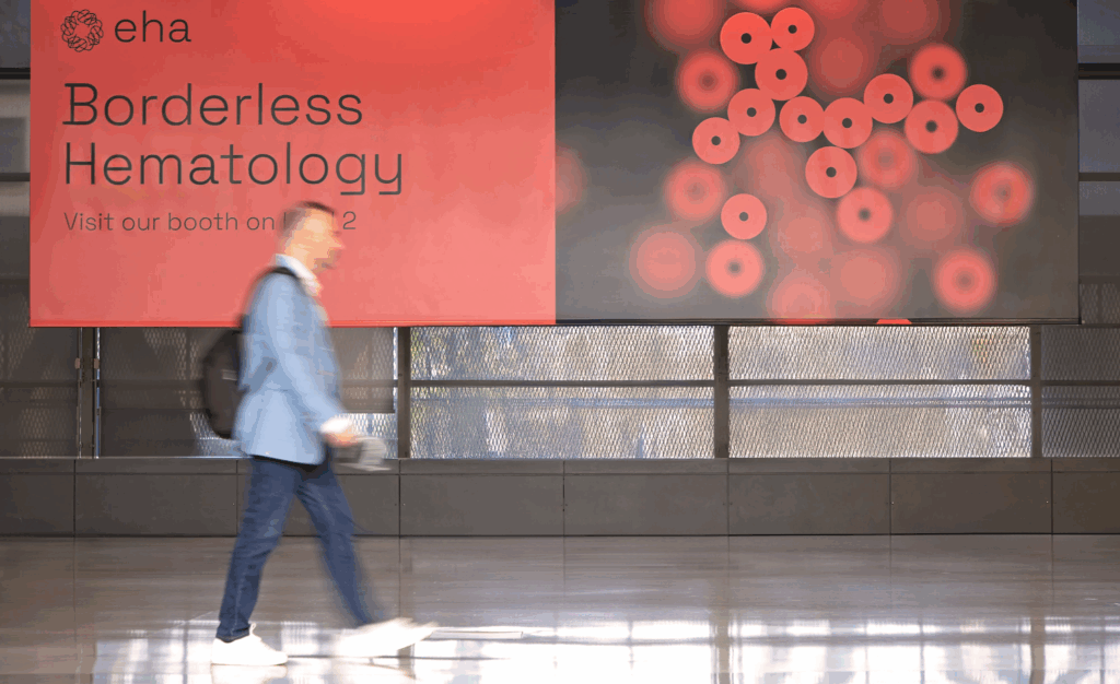 An EHA2025 delegate walking past a large EHA banner. The text of the banner says: 'Borderless Hematology, Visit our booth on level 2.'