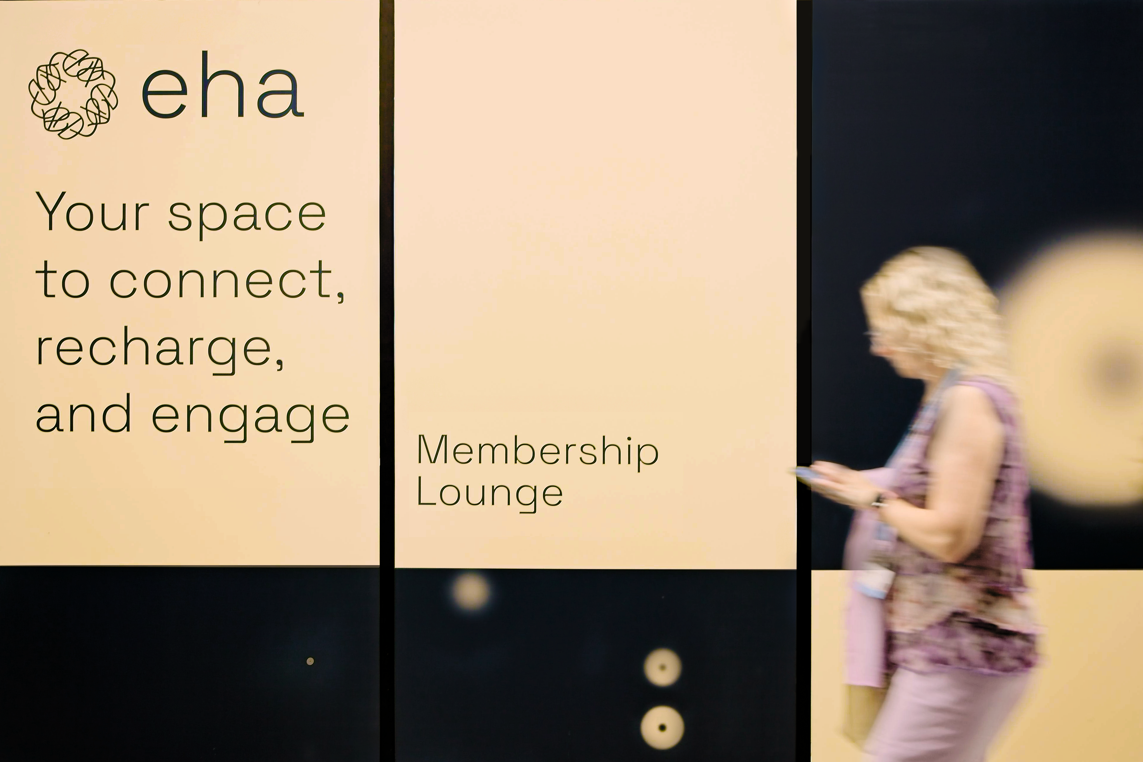 A person walking past the EHA Member Lounge at EHA20205. The text on the left-hand panel says: 'Your space to connect, recharge, and engage.'