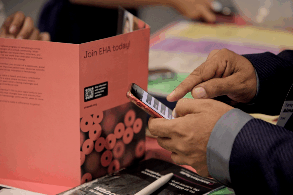 A person looking at the smartphone version of the EHA2025 program. Next to their hand is a 'Join EHA today' leaflet.
