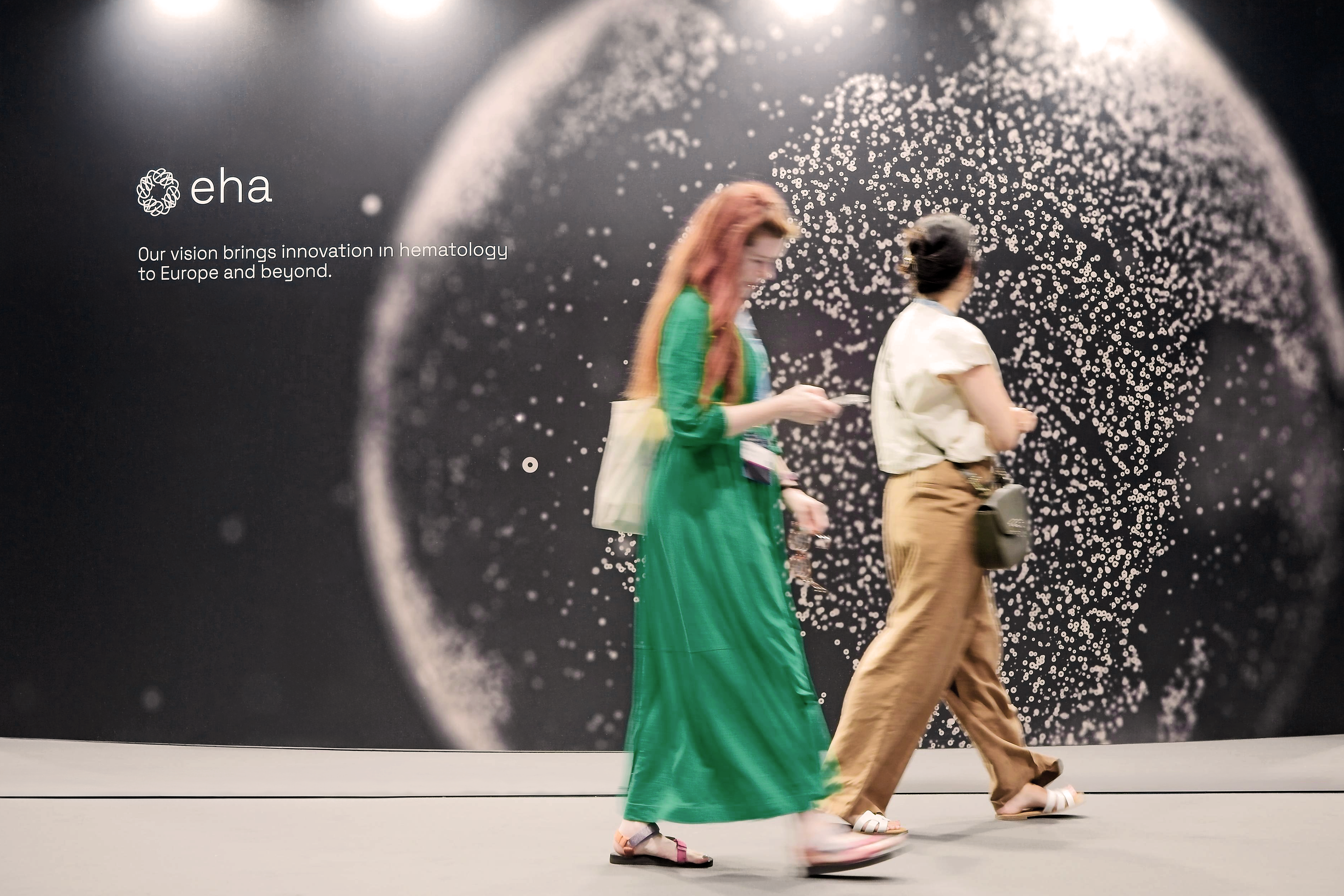 Two EHA2025 delegates walking past a large EHA graphic featuring a globe.