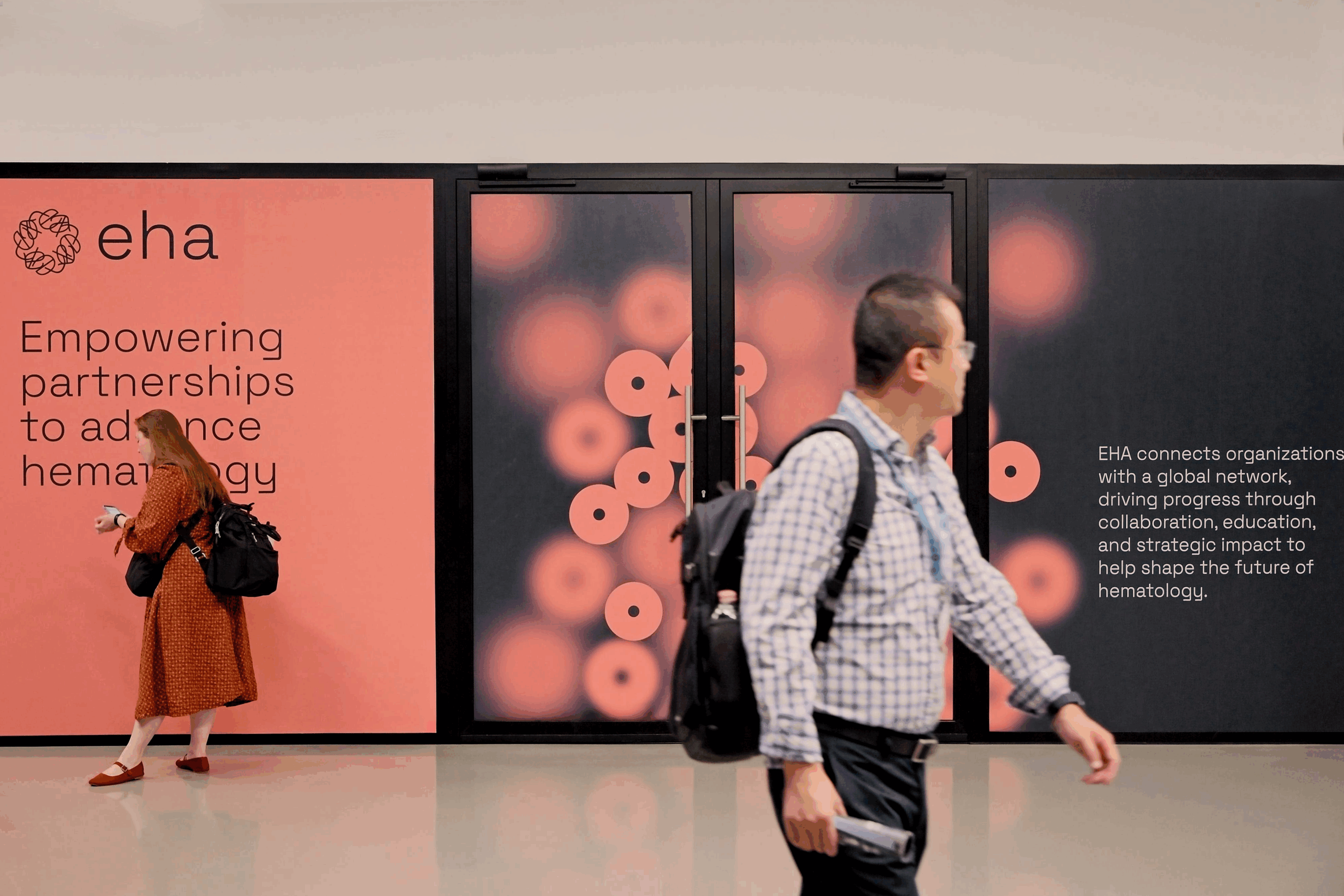 Two delegates walking past some EHA promotional signage that's been placed on doors and walls. The text on the left-hand panel says: 'Empowering partnerships to advance hematology.'